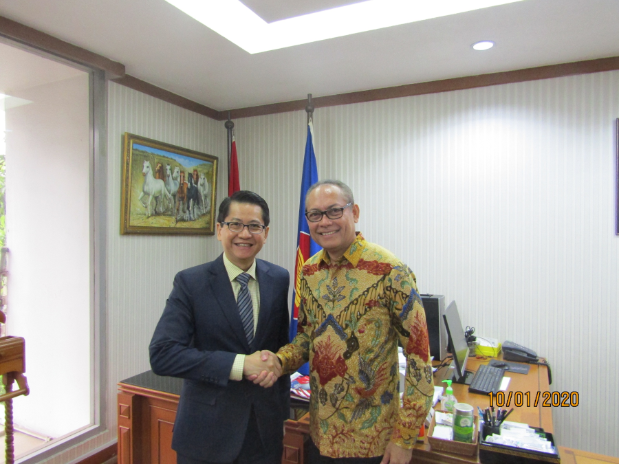 Two men shake hands in an office with flags, artwork, and a desk.